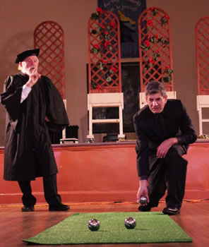 Holofernes in an academic gown and morter board with a finger up to his lips and his left hand on his hips talking as Nathanial kneels on one knee to cast a metal ball down an Astroturf rug where two other balls already sit: in the background, on a stage are trellises of roses on wheeled countertops
