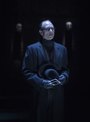 Tyrrell in black overcoat, black turtleneck, wearing glasses and wearing gloves as he holds his black fedora hat in his left hand and two poloroid prints in his right.