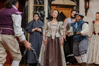 ASC-Comedy_Errors-Cast Antipholus of Ephesus with back to camera and holding his rope as across from him the crowd stares at him: Adriana in greentrimed off-green and brown Elizabethan dress, Pinch in Renaissance violet justice robe and glasses calmly standing with hands clenched at his waist, The courtesan in a bodice, dress, and bushy beard, Dromio of Ephesus in blue vest and cap, blue striped knee pants with white stockings, and Luciana in brown and green patterend Elizabethan dress