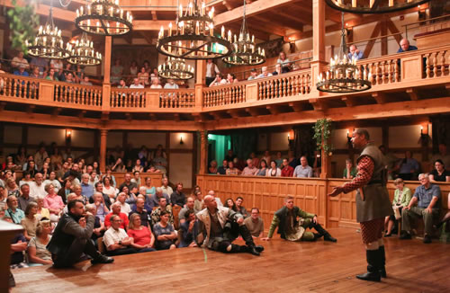 Berowne standing on the stage is in breen short coat over red and gold patterend Elizabetan jacket and knee britches with white stockings and black boots. His hands are outstretched. Sitting at the front of the stage, facing him and backs to the audience, each with an arm on a knee, are Dumaine, Ferdinand, and Longaville. All around (including sitting on stools on the stage) are the audience filling the all-wood, two-tier  theater with more than a dozen iron chandeliers hanging above and flower vines on the posts.