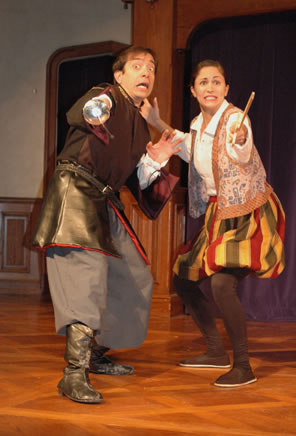 Antipholus wearing brown leather tunic-like jacket, gray pants and brown boots, sword in right hand and left hand up in fearful surprise; Dromio in brocaded best, white shirt, multicolor-striped puffy knee pants with brown stockings, wood sword in left hand and right hand up in fearful surprise