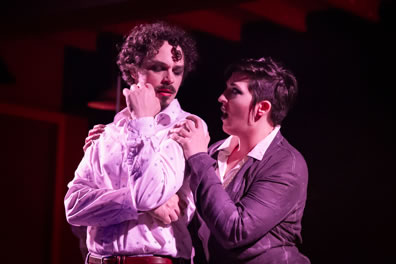 Soranzo in white blousy shirt, right arm crossed over his belly, left bent up with hand on chin as he turns his head to listen to Vasquez with Vasquez in short leather jacket and pants with white shirt and claspping Soranzo's shoulders from behind.