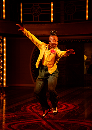 Jester with orange shirt and jacket, carnation in jacket button hole, dark pants, and black and white tap shoes is airborne with outstretched arms as he dances. Photo by Daniel Rader
