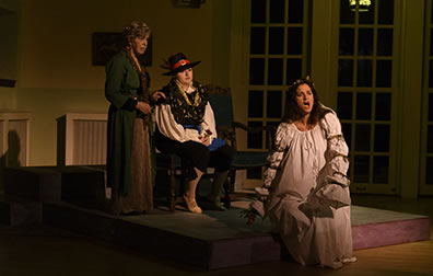 Production photo by Genneth Garrett of Ophelila in a white nightgown sitting on the edge of a platform with Osric sitting on a chair and a waiting woman standing by behind Ophelia.