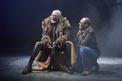 STC-Lear_Page&Fool-1580 Patrick Page as King Lear sits on a case as Michael Milligan as the Fool kneels next to him. Lear is wearing a fur trimmed long coat, brown leather pants and boots and fingerless gloves. The Fool, folded arms against the cold, is wearing a camoflage jacket and blue jeans and glasses.Photo by DJ Corey Photography