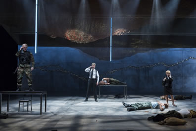 With bodies lying on the stage, the Weird Sisters talk on cell phones, one in combat gear standing on a table to the left, one in white shirt, black pants and shoulder holster standing at the back center, the woman in business suit and skirt standing to the right.