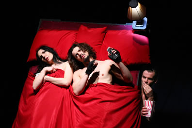 Kate and Petruchio in a bed with red sheets watching porn TV as Grumio, eating popcorn, peers out from under the bed